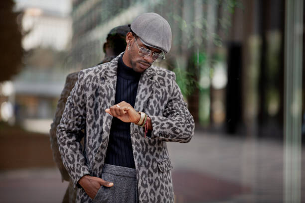 Handsome black man in stylish clothing looking at wristwatch while standing near glass wall in city. He is wearing trendy cap, coat and glasses stock photo