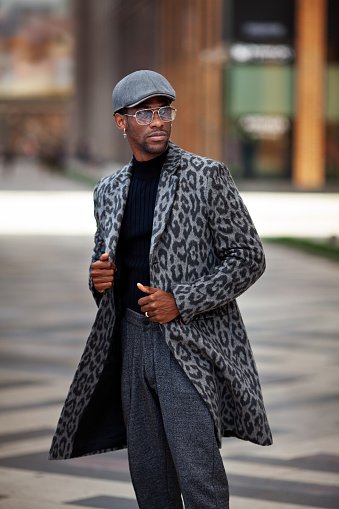 Portrait African American young male in trendy outfit coat with animal print, turtleneck, cap, pants, and glasses, standing on city street. Handsome man, fashion style.