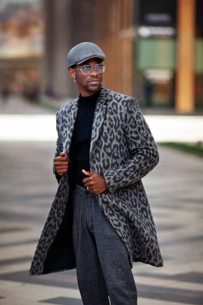portrait african american young male in trendy outfit coat with animal print, turtleneck, cap, pants, and glasses, standing on city street. handsome man, fashion style - fashion men fashion model male imagens e fotografias de stock