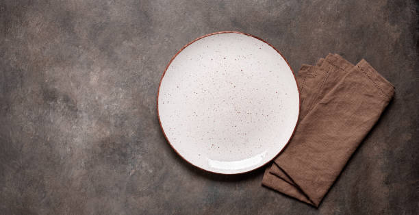 istock White plate with a brown napkin on a brown rustic background. Top view, flat lay, copy space. Banner. 1672421570