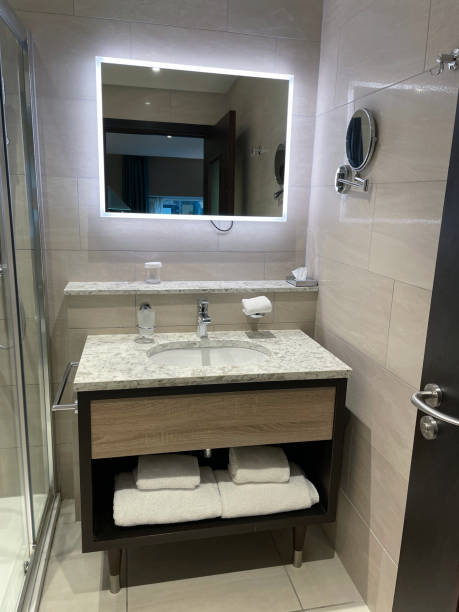 istock Image folded, white towels stacked on wooden shelving unit under granite bathroom counter, bathroom sink with chrome monobloc mixer tap, granite shelf under wall mirror, glass shower cubicle, tiled flooring, focus on foreground 1672434311