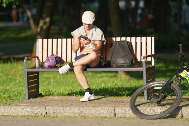Moscow, Russia - May 27, 2023: Man sits on the bench in the park. He is rests in the sunny warm day. He using smartphone, looking at the screen and smiling. His bike is nearby. Street photography