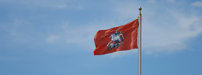 Red flag flutters. It's coat of arms of Moscow is drawn on flag. Saint George and the Dragon. Waving flag in front of the blue sky in sunny summer day. Panoramic image. Close up image