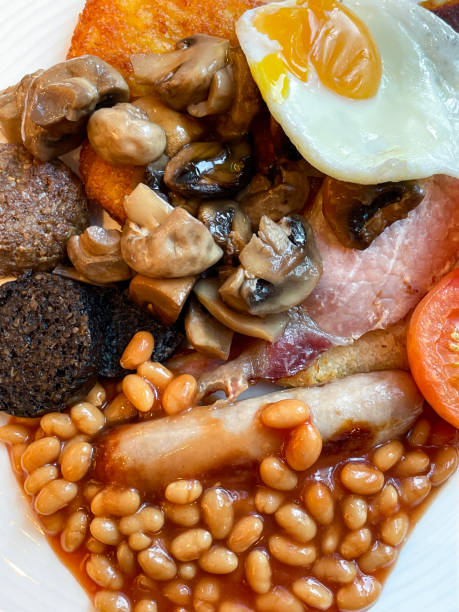 Stock photo showing close-up, elevated view of a white plate containing ingredients of a Full English breakfast with heap of baked beans, sunny-side-up fried egg, pile of mushrooms, sausage, grilled tomatoes, black pudding, bacon rasher and two hash browns. imagen de fotograma completo de ingredientes de desayuno fritos ingleses completos, montón de frijoles horneados, huevo frito con el lado soleado, pila de champiñones, salchichas, tomates a la parrilla, morcilla, rasher de tocino y dos papas fritas en pl