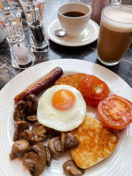 Stock photo showing close-up, elevated view of a white plate containing ingredients of a Full English breakfast with sunny-side-up fried egg, pile of mushrooms, sausage, grilled tomatoes and two hash browns. imagen de ingredientes de desayuno fritos ingleses completos, huevo frito con el lado soleado, pila de champiñones, salchichas, tomates a la parrilla y dos papas fritas en plato blanco, configuración de la mesa del restaurante, vista elevada