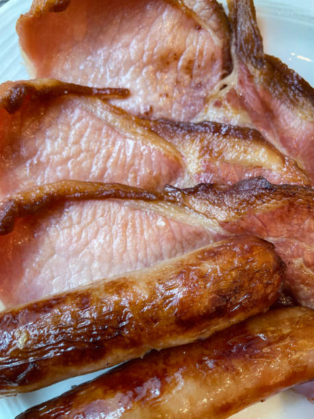 istock Full frame image of Full English fried breakfast ingredients, three bacon rashers and two sausages on white plate, elevated view 1672479035