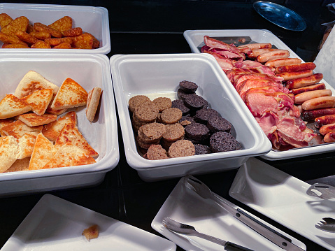 Stock photo showing elevated view of a Full English fried breakfast self service buffet, with large white ceramic dishes filled with rows of bacon rashers, grilled sausages, white and black pudding, potato farls (potato cake triangles) and hash browns on a hotplate food bar. The breakfast dishes are being kept warm beneath heat lamps so that guests can help themselves with the tongs provided.