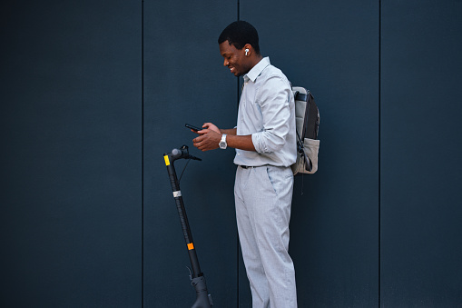 A side view of a smiling African-American male with a scooter listening to music using an app on his smartphone while being outdoors.