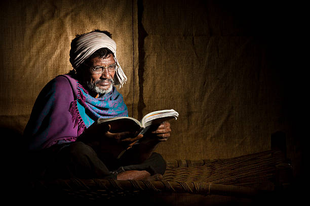 Senior, bespectacled, rural Indian man reading a book This conceptual image is based on portraying literacy in rural India. A senior, bespectacled, rural Indian man is reading a book while sitting on a traditional woven jute cot called a Charpai. The old man has a white beard and gray moustache and is wearing a blue shawl and white turban. A jute curtain hangs in the background. This low-key portrait composed in a square format allows for ample copy space.  human hand traditional culture india ethnic stock pictures, royalty-free photos & images