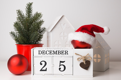 Red christmas ball in santa hat on calendar with December 25 date,some gift bags around,christmas tree and decorative houses.