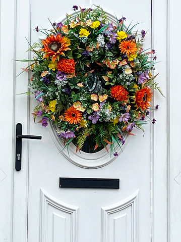 Stock photo showing a white, front door with an artificial, summer wreath decoration of green foliage and orange and purple flowers including gerbera.