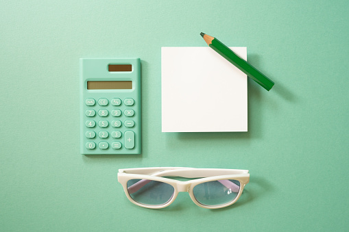 Green calculator and white memo note pad, colored pencil, eyeglasses on green background. top view