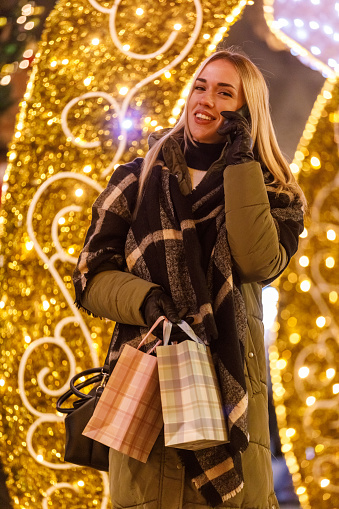 Portrait of joyful young blond woman standing by the glistening Christmas lights on the city street, holding gift and shopping bags, talking on the phone while waiting for a friend to meet.