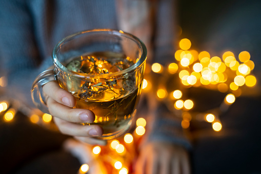 Herbal tea in a glass mug glowing in the reflection of the illuminated fairy lights held by a hand wearing a jumper