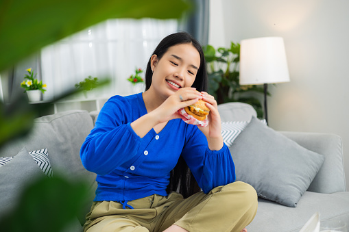 Portrait of young asian woman sitting on sofa and holding a burger to eat while rest in living room,  She Indulging in delicious burger at home, junk food delivery concept