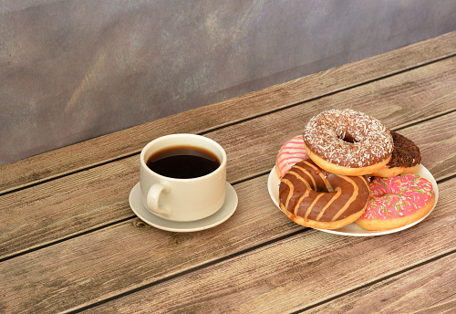 A cup of hot black coffee on a saucer and a plate with a bunch of fresh donuts in multi-colored glaze on a wooden table against a gray wall. Close-up.