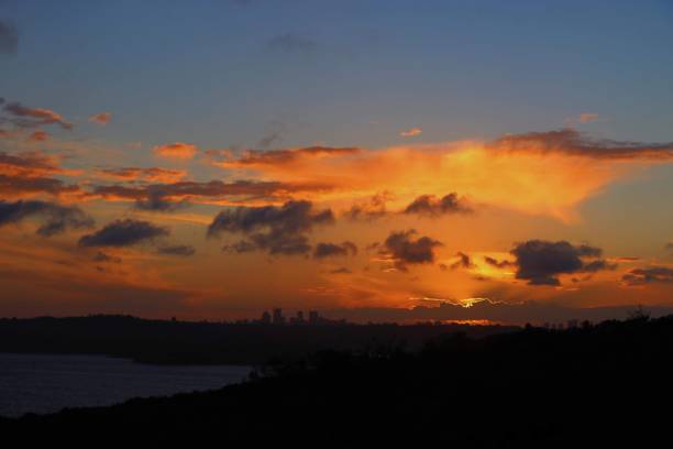 A profile of Sydney from the central business district on the left, the Sydney Harbour Bridge in the centre, and North Sydney on the right. The sun had set behind the skyscrapers on the right horizon, where the largest area of yellow appears. A bank of clouds is visible on the horizon. This image was taken from Dudley Page Reserve, Dover Heights on a windy, cold and sunny afternoon about 30 minutes after sunset on 24 May 2025.