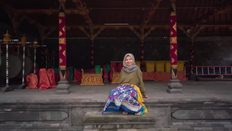 Female with hijab and sarong sitting gracefully, with traditional instruments background