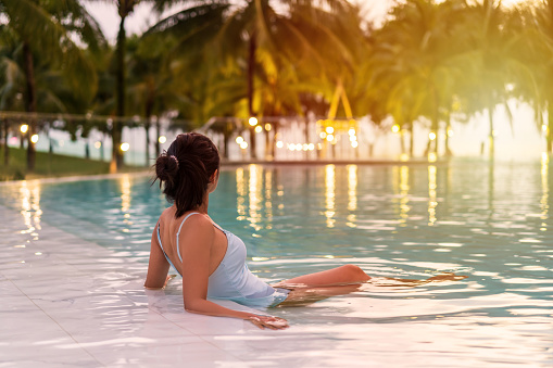 Young woman traveler relaxing and enjoying the sunset by a tropical resort beachfront pool while traveling for summer vacation