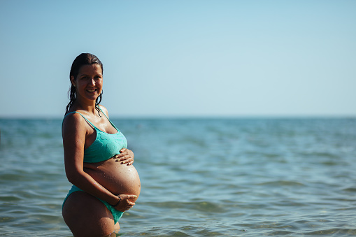 Photo of a eight month pregnant woman with hands on her abdomen at the seaside