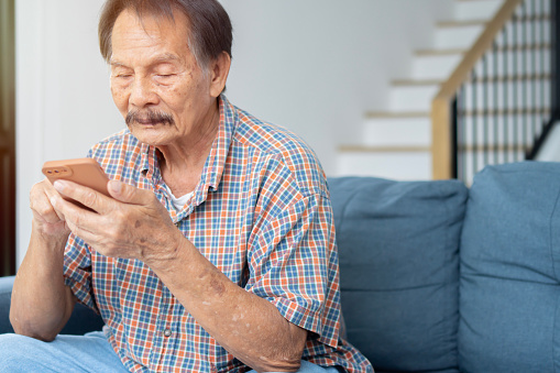 Portrait photo of engaged elderly Asian man holding phone in two hands while relaxing on sofa at home.  Technology concept.