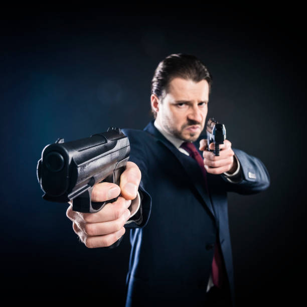 Dangerous man in suit is aiming with two guns, assassin, killer, mafia. Studio shot, dark background, square.