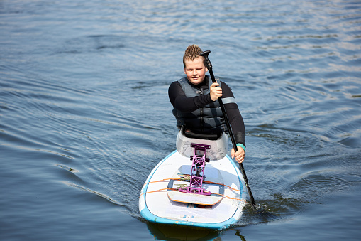 Full length portrait of smiling young boy with disability enjoying paddle boarding on water using adaptive sports equipment, copy space