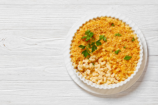 close-up baked mac and cheese sprinkled with panko breadcrumbs and fresh parsley in baking dish on white wooden table, horizontal view, flat lay, free space
