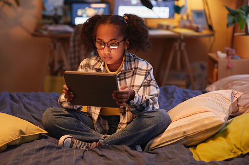 Full length portrait of teen black girl with pigtails using digital tablet while sitting on bed at home in cozy warm light, copy space