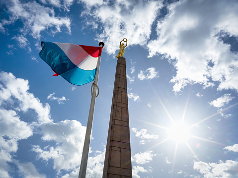 Luxembourg War Memorial Gëlle Fra (Golden Lady) - Monument of Remembrance  (Monument du Souvenir) and Luxembourg National Flag blowing in the wind against blue summer sky, backlit from sun star sun. High-res 102 MPixel Hasselblad X2D Shot. Iconic War Memorial - Obelisk in downtown Luxembourg City. Luxembourg, Benelux, Europe