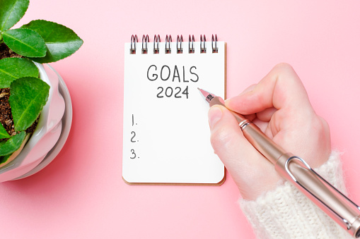 Female hand writes in a paper notebook goals 2024 close up on a pink background.