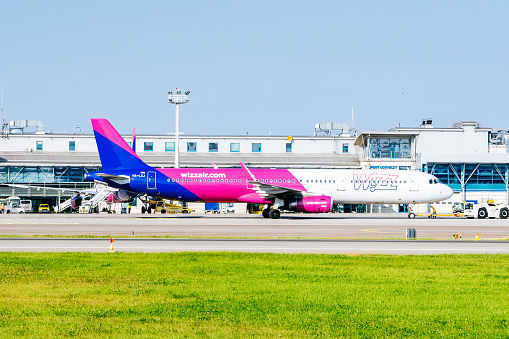 An Wizz Air Airbus A320 is taxiing to takeoff from the Lech Walesa Airport in Gdansk, Poland. The airplane has a purple and blue color scheme and a logo on the tail. The airplane is moving on the tarmac with its engines running. The airport terminal building and other airplanes are visible in the background. The sky is blue and the grass is green, creating a contrast with the colorful airplane.