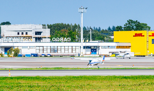 A white and blue propeller plane is taxiing on the runway of the Lech Walesa Airport in Gdansk, Poland. The airport buildings, including a control tower and a yellow DHL cargo building, are visible in the background. The runway is concrete and has several markings on it. The sky is blue and there are a few clouds in the distance. The image shows the plane preparing for takeoff and the activity of the airport.