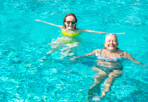 Couple of two mature women friends having fun floating in outdoor swimming pool under the sun. Vacation, healthy lifestyle and relax concept