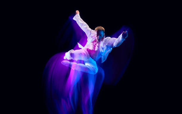 istock Dynamic image of stylish young man in white clothes humping against black studio background in neon with mixed lights effect 1673540861