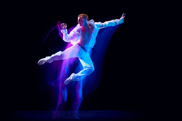 istock Dynamic image of stylish young man in white clothes humping against black studio background in neon with mixed lights effect 1673540870