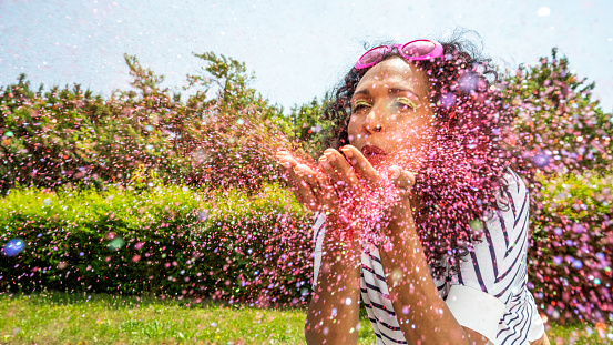 Mature woman blowing glitter confetti against sky.