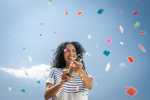 Smiling mature woman exploding confetti from party popper during event.