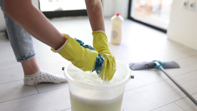 Cleaning lady in rubber gloves squeezes rag with a mop over bucket in room