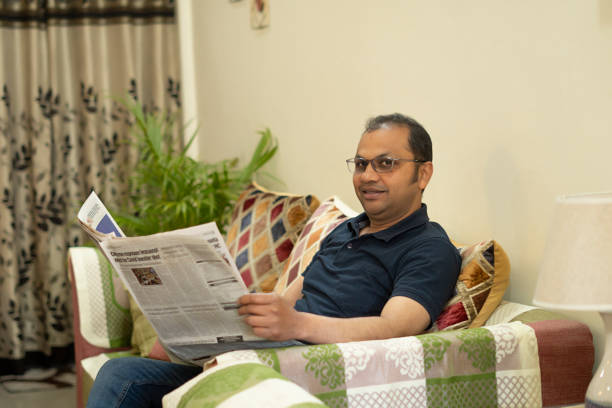 istock Young man reading newspaper sitting on sofa at home 1673548330