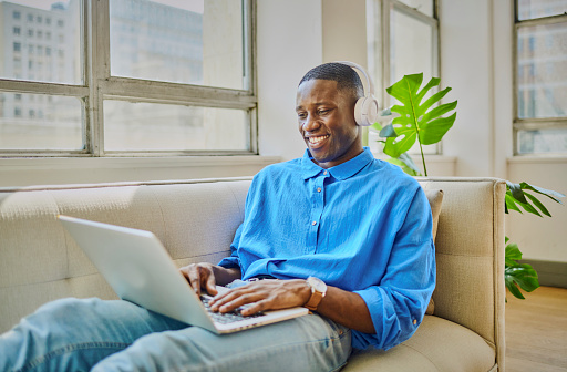 young man smiling at his laptop happy on the sofa using his headphones working remotely.