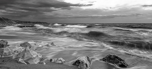 Black and white panoramic seascape with waves flowing between rocks.