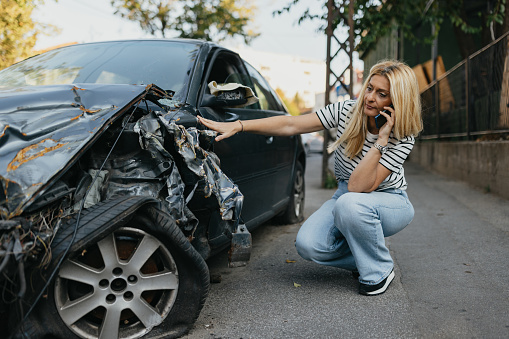 A young adult woman, the sole occupant at a car accident scene, leans against the railing by the road. She holds her mobile phone, gesturing with frustration and anxiety as she navigates the emotional stress caused by the accident.