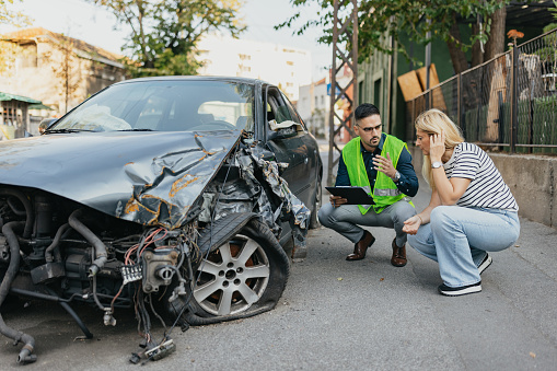 A woman in her late 40s, visibly worried and stressed, stands beside her damaged car after a car accident. She's using her mobile phone to speak with her insurance agent, a young man, as they navigate the process of reporting and seeking assistance.