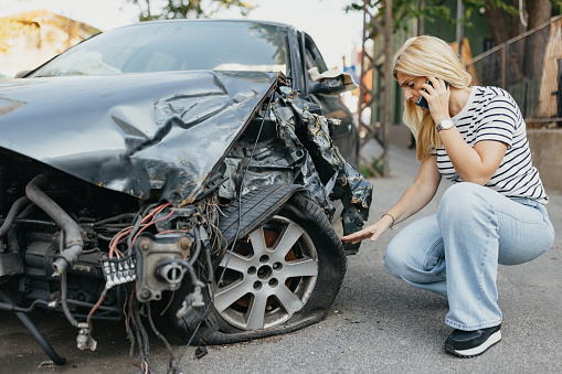 An adult woman stands outdoors, holding her mobile phone while looking down with concern. In the face of car damage and inconvenience due to a crash, she relies on wireless technology to communicate, listen for help, and overcome the imperfections of an unexpected roadside situation