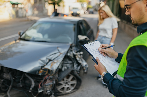 A young insurance agent examines the damaged car's front view on the roadside. He provides expert assistance to the mature woman involved in the accident, helping her fill out necessary forms to report the incident.