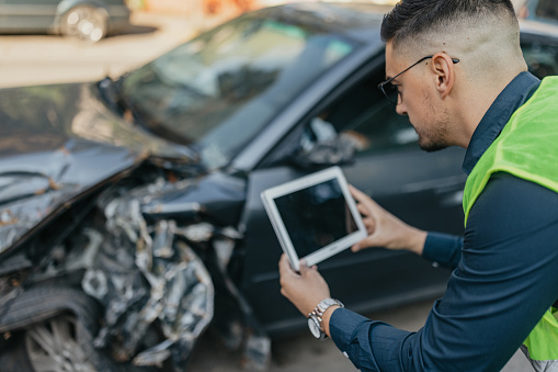 An insurance agent, a man in his early 30s, stands outdoors as he carefully documents the damaged car for an insurance claim. With precision and professionalism, he works to support the car owner during this challenging time of misfortune, ensuring a smooth claims process.