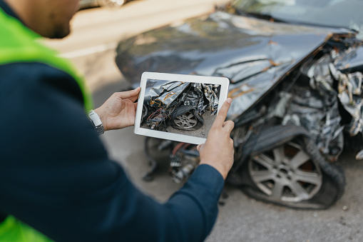 A male insurance agent in his early 30s stands outdoors, expertly examining a damaged car after a recent traffic accident. With a digital tablet in hand, he meticulously assesses the extent of the dented bumper and broken vehicle part, demonstrating his expertise in managing car insurance claims.