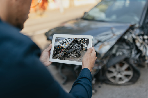 An insurance inspector, a man in his early 30s, employs his expertise while examining a damaged car outdoors. With a digital tablet in hand, he meticulously assesses the dented car bodywork and broken bumper, ensuring a thorough and efficient car insurance claim process.
