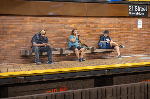 21th Street Queensbridge Subway Station, Long Island City, Queens, New York, USA - August 23th 2023: Three people sitting on a bench while waiting for the subway train